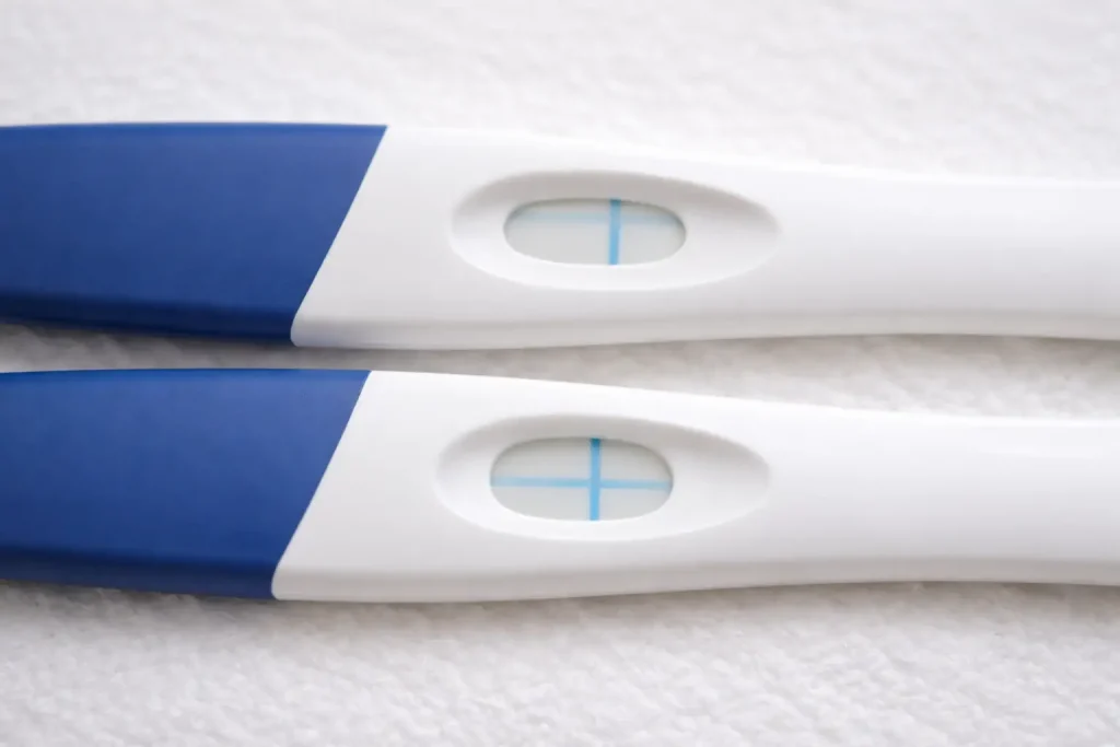 : Gros plan sur deux fenêtres de résultat de tests de grossesse sur fond clair, l’une montrant une ligne horizontale et l’autre une ligne verticale (ou un “plus”), sans texte ni marque visible